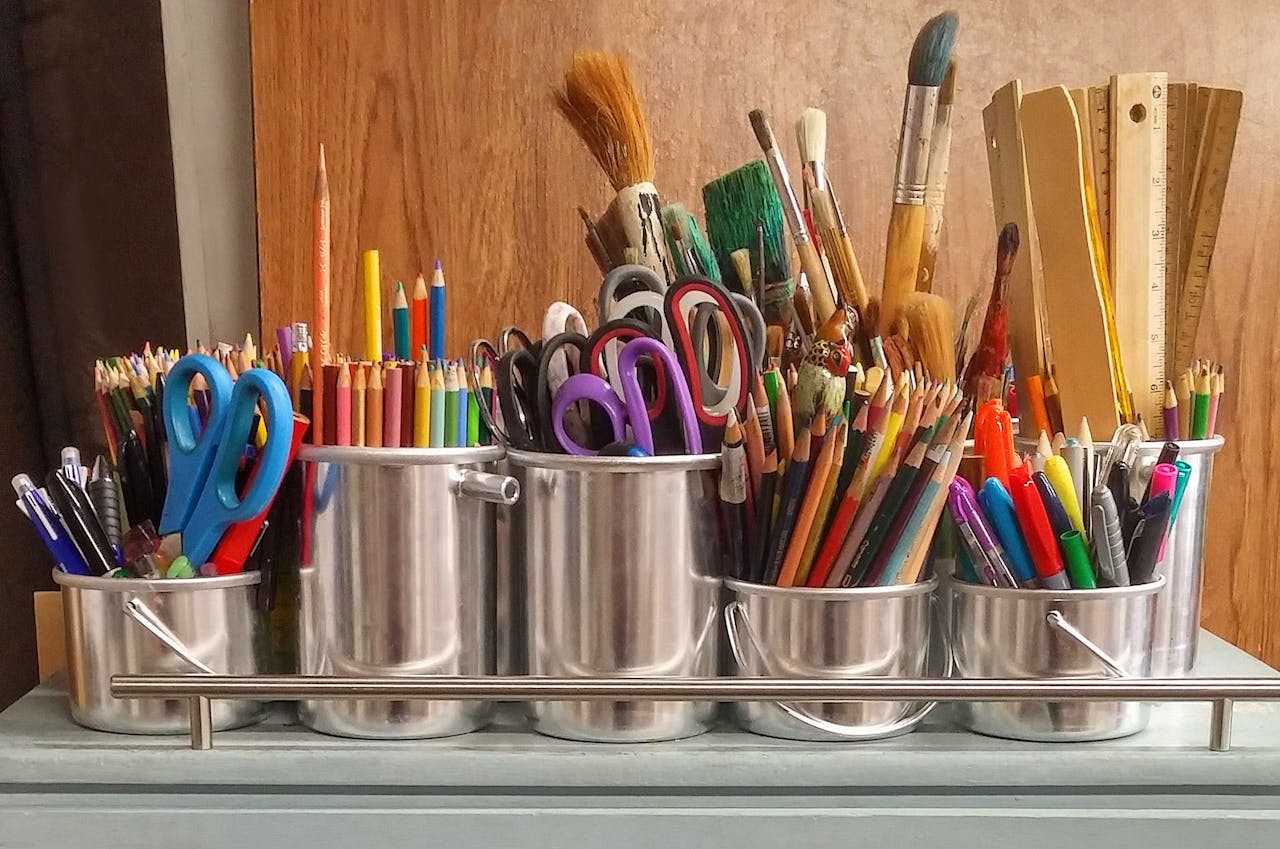 heros-img A diverse collection of art supplies in metallic buckets on a wooden table.